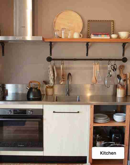 Cozy kitchen with stainless steel countertop, sink, kettle, hanging utensils, and open shelf with dishes and books above the counter.